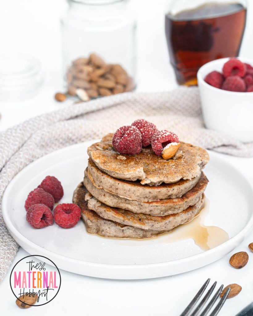 Five almond flour pancakes on a plate with syrup and berries. Towel in background. 