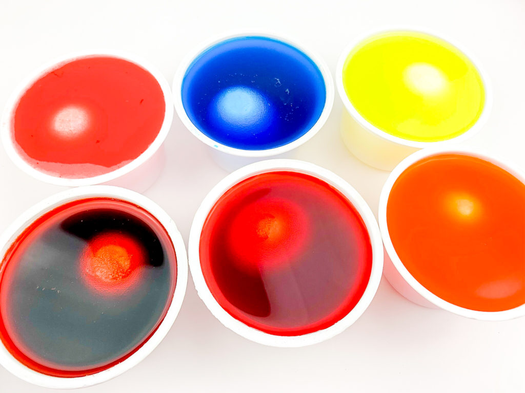six eggs dyeing in recycled yogurt cups filled with different colors of Kool-Aid and water mix