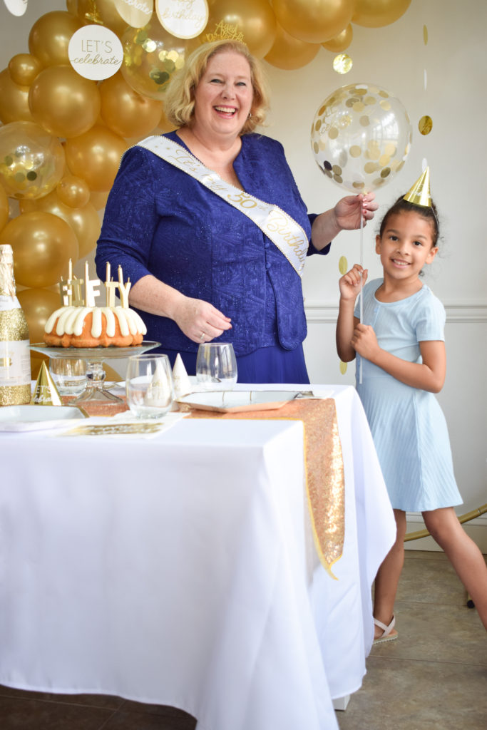 50th birthday party with a woman wearing a blue dress, holding a clear balloon on a stick filled with gold confetti. She is handing the balloon to her 6 year old granddaughter while standing behind a table set with a birthday cake, paper plates, napkins, gold party hats, drinking glasses, a 50 number sign, and a confetti popper bottle.