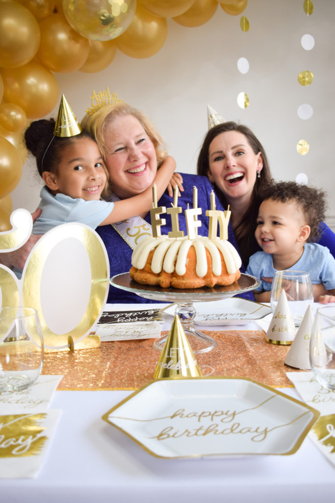 A family including a mother, her daughter, and her two grandchildren celebrating her birthday. Granddaughter hugging her grandmother. A cake with candles that say "Fifty" on a glass stand surrounded by place settings with paper plates, party hats, drinking glasses and a 50 number sign. Gold balloons in the background.