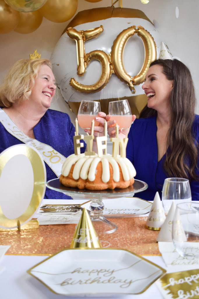 50th birthday party with a woman wearing a blue dress, sitting at a table with her daughter in a blue dress, holding glasses while giving birthday cheers. Table set with a birthday cake, paper plates, napkins, gold party hats, drinking glasses, and a "50" number sign with a gold balloon arch and "50" golden age themed balloon in the background.