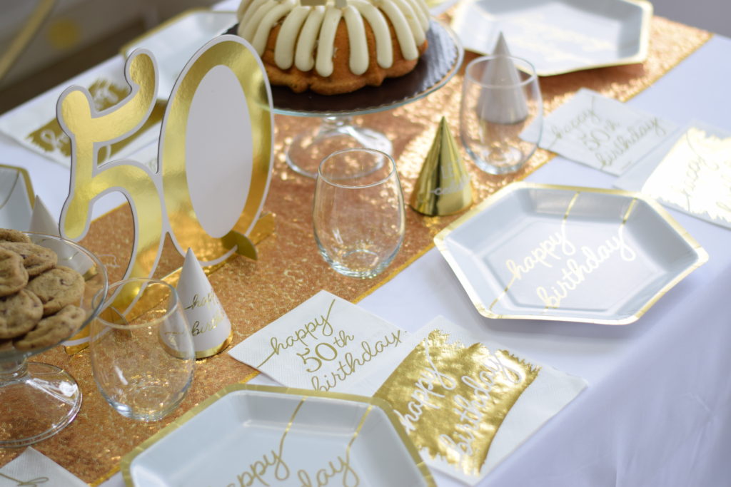 Table covered with white tablecloth, set with place settings including paper plates, napkins, party hats, drinking glasses, gold glitter table runner, a birthday cake, gold "50" number sign, and a glass stand holding chocolate chip cookies.