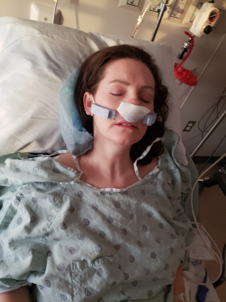 Woman laying in a hospital bed after septoplasty wearing a nasal dressing holder with gauze and a hospital gown.