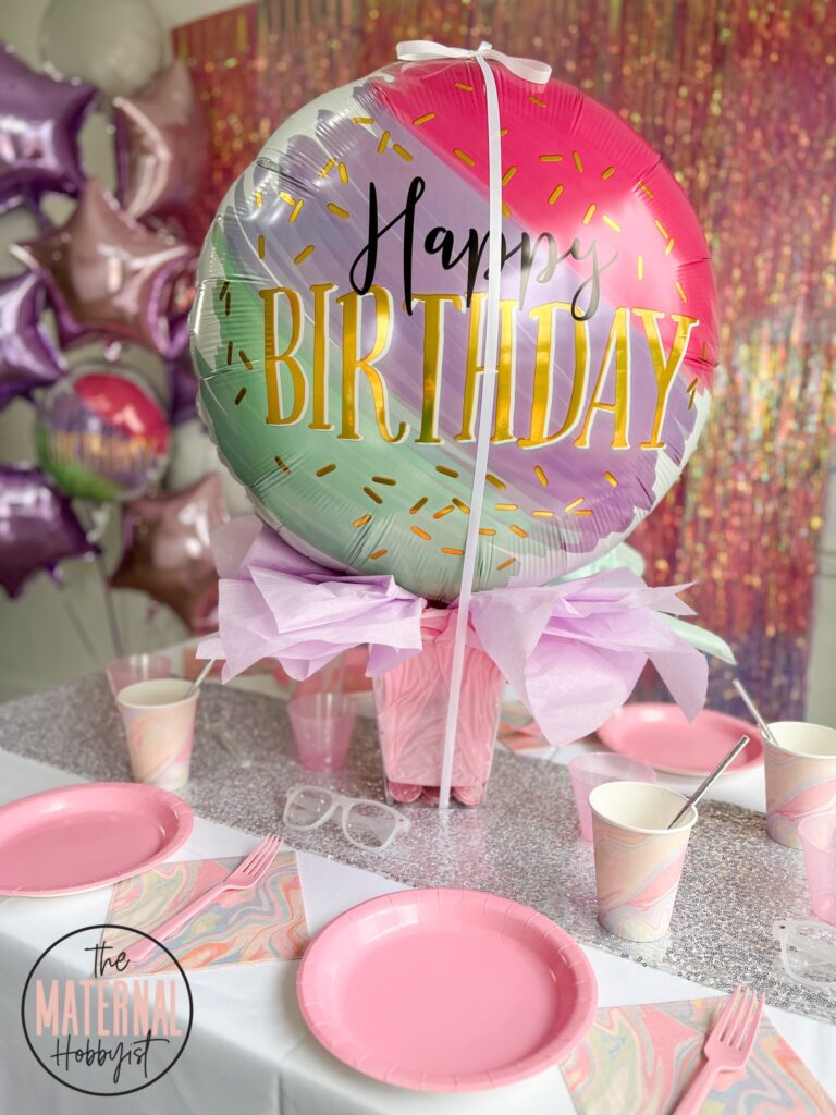 giant helium happy birthday watercolor style, pastel colored balloon tied with a white ribbon sitting on a clear plastic container filled with pastel tissue paper on a table. White table cloth with a silver sequin table runner set with four place settings with pink plates, and pastel marble colored napkins and cups with silver paper straws. Birthday bouquet of balloons in the background with a shimmer pink and purple backdrop.