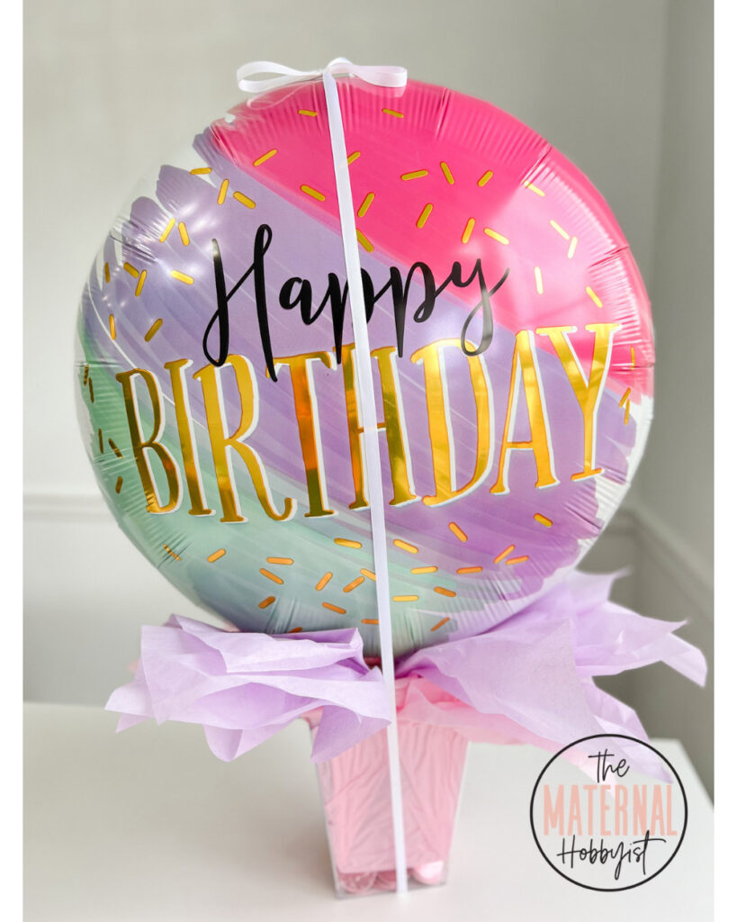DIY money train balloon surprise using a giant helium happy birthday balloon tied with ribbon to a plastic container filled with purple and pink tissue paper.