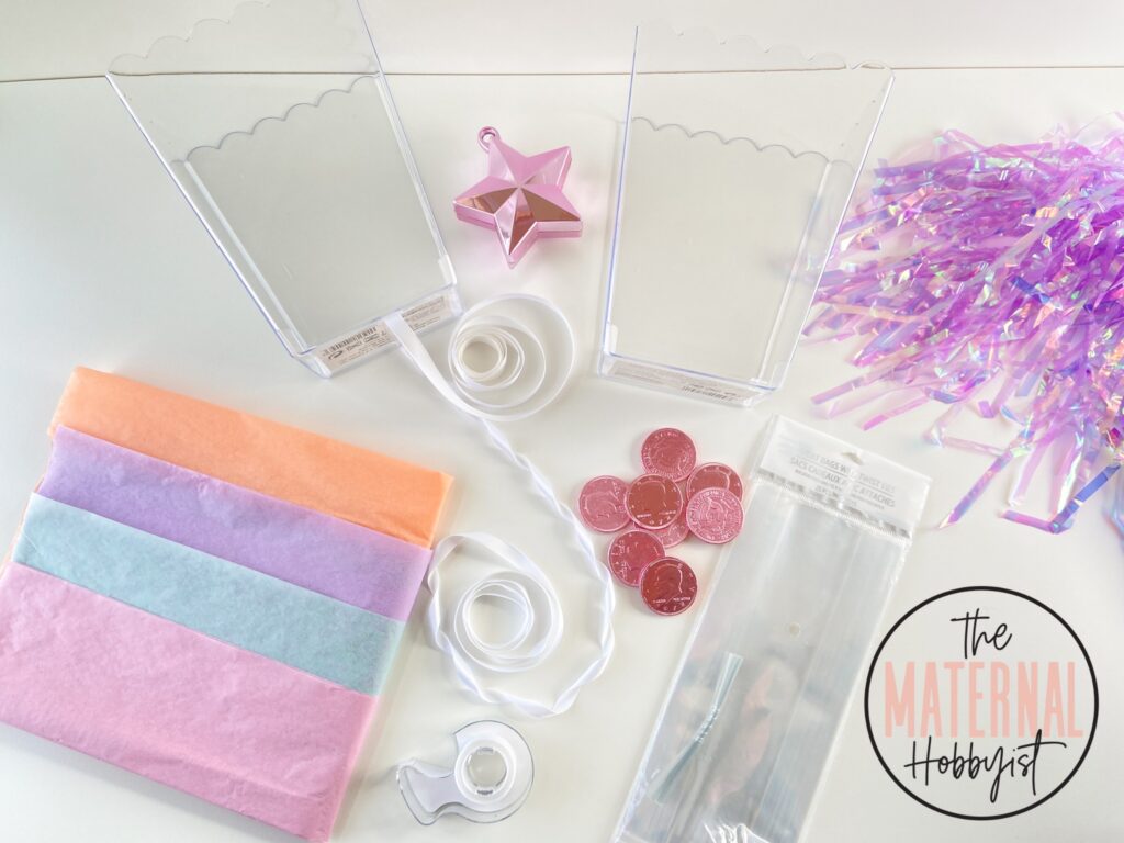 DIY Money Train Balloon surprise supplies including pastel tissue paper, two clear plastic containers, one pink star balloon weight, white ribbon, pink chocolate coins, small clear plastic bags and purple decor.