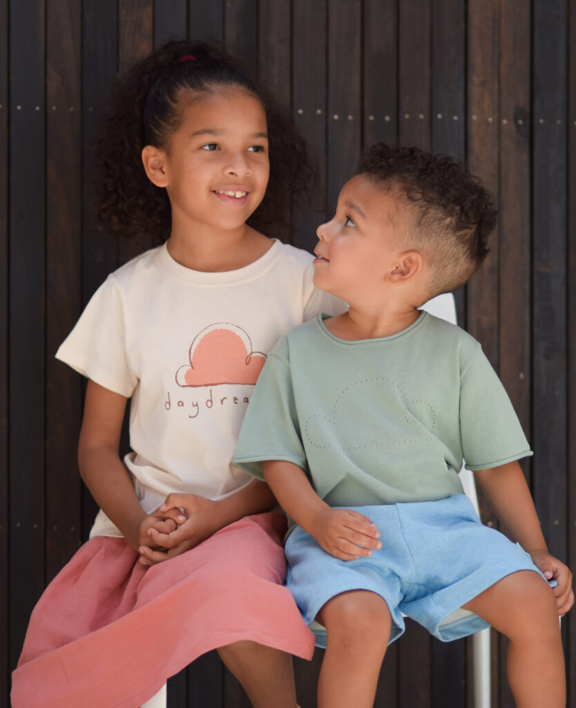 Girl wearing Petite Revery white shirt with pink cotton skirt. Younger boy sitting next to her wearing Petite Revery green cloud shirt with blue cotton shorts.