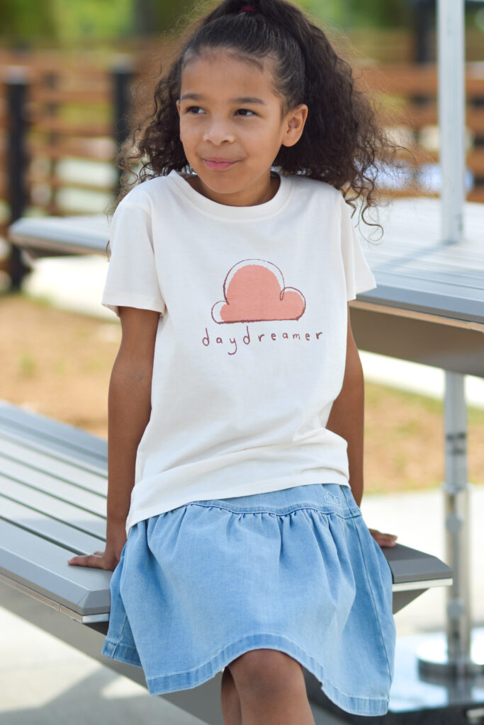 Girl sitting while wearing Petite Revery "Daydreamer" shirt in cream color with denim skirt.