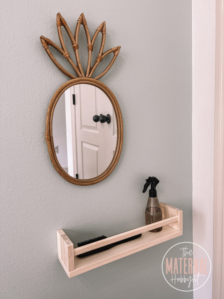 Pineapple shaped mirror on the wall above a wall shelf holding a brush and a spray bottle of water.