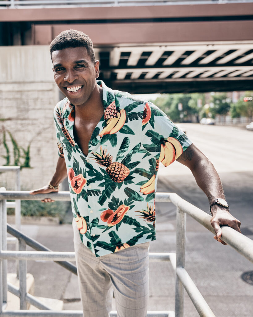 African American male named Kenneth hill wearing a Hawaiian shirt with pineapples, fruit and palm leaves and dress pants standing in the corner of a railing with a smile.