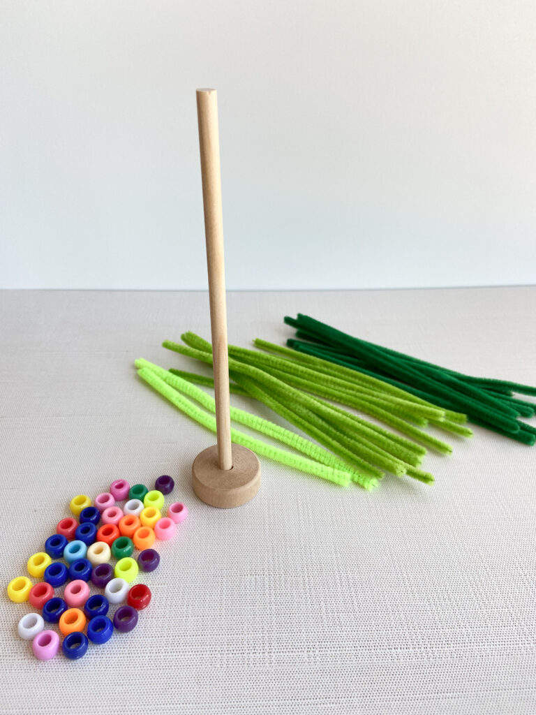 wooden dowel rod stuck into a wooden wheel next to colorful pony beads and green pipe cleaners on a table.