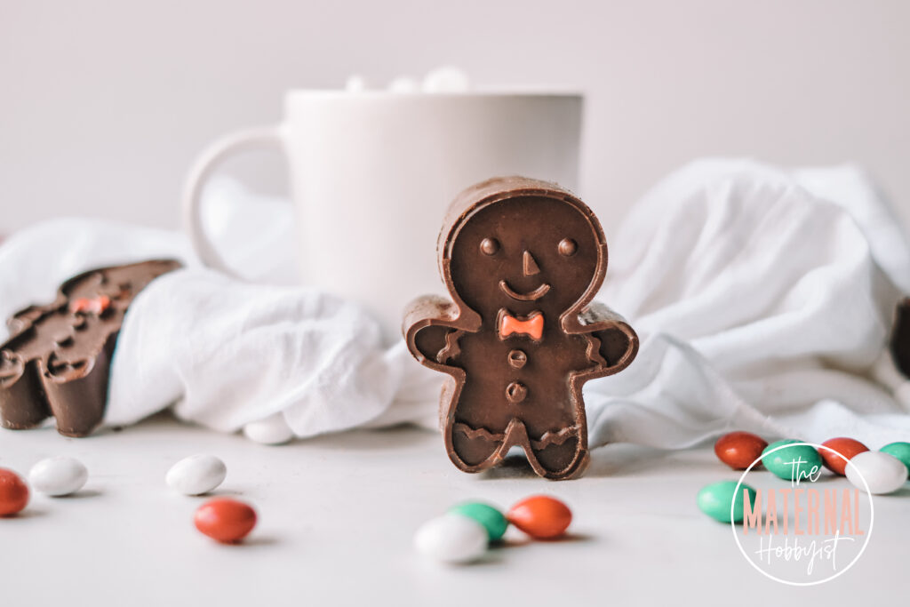 Gingerbread shaped hot cocoa bomb with a red chocolate bow tie standing on a white counter surrounded by red, green and white M&M's.